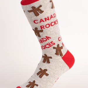 Może przedstawiać: Szare skarpetki z czerwonymi akcentami na palcach i mankietach. Skarpetki zdobią brązowe symbole Inukshuk i czerwony napis "CANADA ROCKS". Zabawny i patriotyczny dodatek.