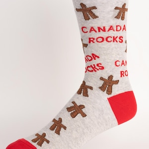 Puede incluir: Un calcetín gris claro con puntera, talón y puño rojos. El calcetín presenta símbolos Inukshuk marrones y las palabras "CANADA ROCKS" en rojo. El calcetín está diseñado para ser usado en el pie.