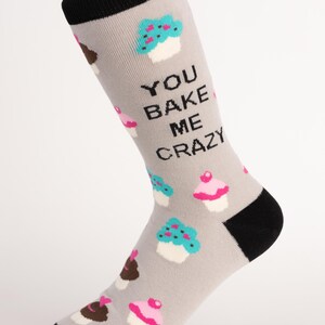 May include: Grey socks with a black heel, toe, and cuff. The socks feature a pattern of cupcakes in various colours and the text "YOU BAKE ME CRAZY".