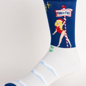 Könnte beinhalten: Novelty-Socke mit blauem Bündchen und Zehen. Die Socke zeigt ein Cartoon-Bild einer Frau mit Weihnachtsmütze, die ein Schild mit der Aufschrift "North Pole" hält. Das Design der Socke umfasst einen weißen Fuß mit blauen Akzenten.