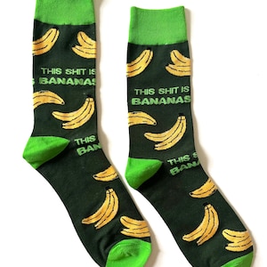 May include: Novelty socks with a dark green base, decorated with yellow banana motifs and the white text "THIS SHIT IS BANANAS". The toe and cuff of the socks are bright green.
