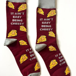 May include: Burgundy socks with grey heels and cuffs, decorated with yellow cheese wedges and the text "IT AIN'T EASY BEING CHEESY". A playful and humorous design for a fun accessory.