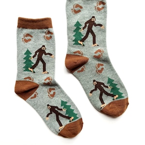 Può includere: Un paio di calzini grigi con polsino marrone. I calzini presentano un motivo ripetuto di alberi verdi, impronte marroni e figure marroni di Bigfoot che camminano.