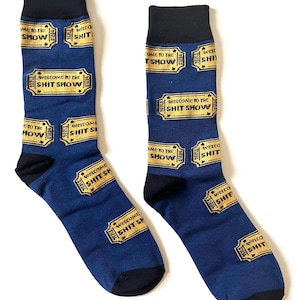 Puede incluir: Un par de calcetines azules con talones y dedos negros. Los calcetines presentan un patrón repetido de talones de boletos dorados que dicen "WELCOME TO THE SHIT SHOW". El texto "COME TO THE SHOW" también es visible.