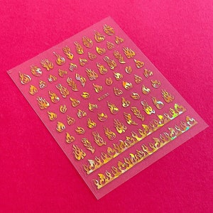 May include: A sheet of clear nail decals with gold holographic flame designs. The sheet includes a variety of flame sizes and shapes.