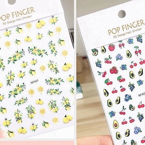 May include: Two sheets of 3D nail stickers. One sheet features lemons and leaves on a white background. The other sheet features cherries, blueberries, raspberries, and avocados on a white background. Both sheets have the text "POP FINGER 3D Design Nail Sticker" and a product code.