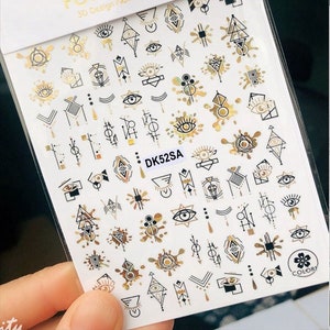 May include: A sheet of gold and black nail stickers with geometric designs, including eyes, triangles, and abstract shapes. The sheet is labeled "POP FINGER 3D Design Nail Sticker" and "DK52SA".