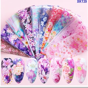 Op de afbeelding: Een set van 16 nagelkunst-transferfolies met bloemmotieven. De folies zijn in een waaier vorm gerangschikt en tonen een verscheidenheid aan kleuren, waaronder roze, paars, blauw en groen. De folies zijn ontworpen om te worden gebruikt met nagellak om unieke en ingewikkelde nagelontwerpen te creëren.