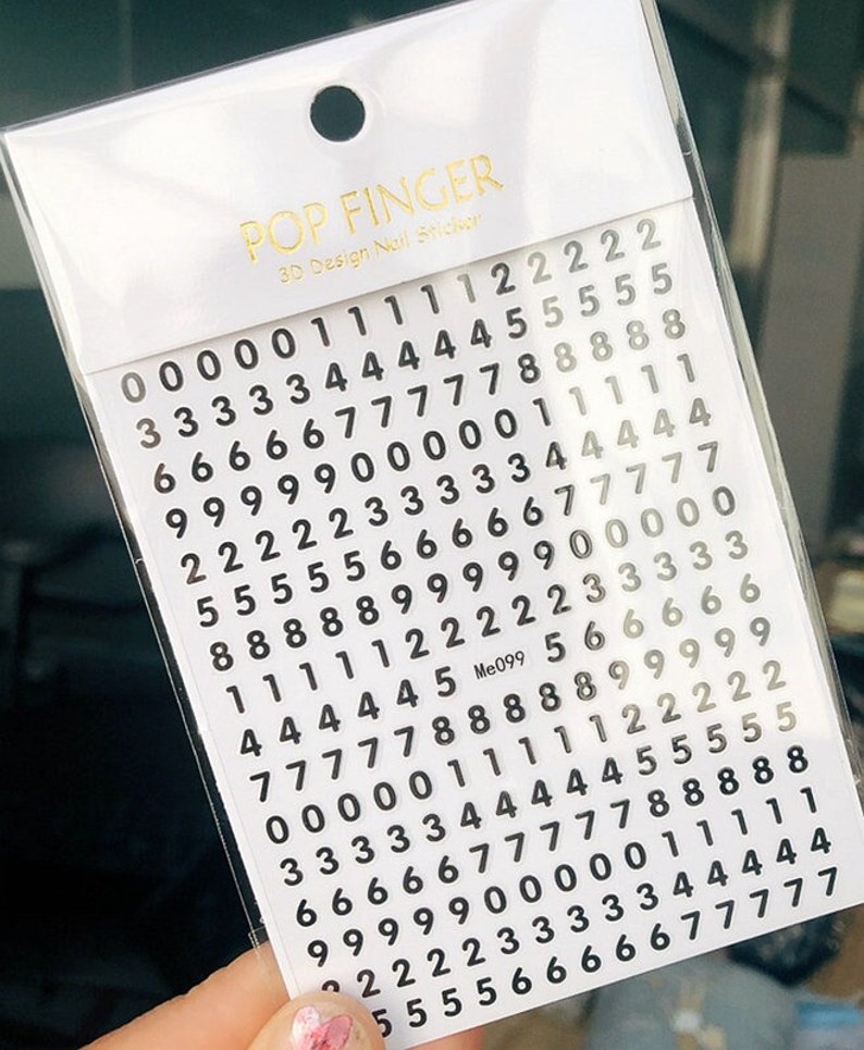 May include: A sheet of black number stickers on a white background. The numbers range from 0 to 9 and are arranged in rows. The sheet is labeled "POP FINGER 3D Design Nail Sticker" and "Me099".