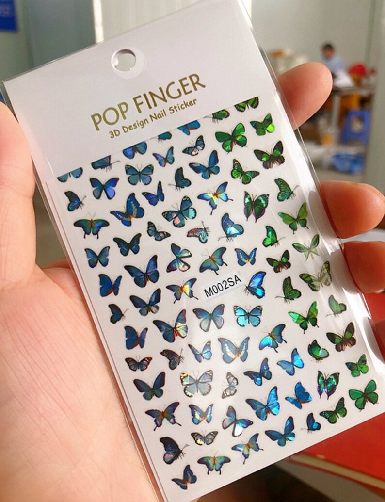 May include: A sheet of nail stickers with a design of iridescent blue and green butterflies. The sheet is labeled "POP FINGER 3D Design Nail Sticker" and "M002SA".