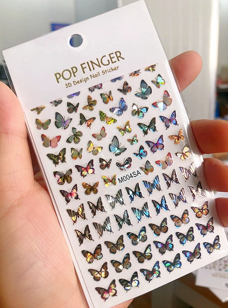 May include: A sheet of iridescent butterfly nail stickers. The stickers are arranged in rows and feature a variety of butterfly designs, including butterflies with wings that are striped, spotted, and solid colors. The sheet is labeled "POP FINGER 3D Design Nail Sticker" and "M004SA".
