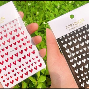 Op de afbeelding: Twee verpakkingen met 3D nagel stickers met een hartjes ontwerp. E&eacute;n verpakking heeft rode hartjes op een witte achtergrond en de andere heeft witte hartjes op een zwarte achtergrond. De verpakkingen zijn gelabeld met "POP FINGER 3D Design Nail Sticker".