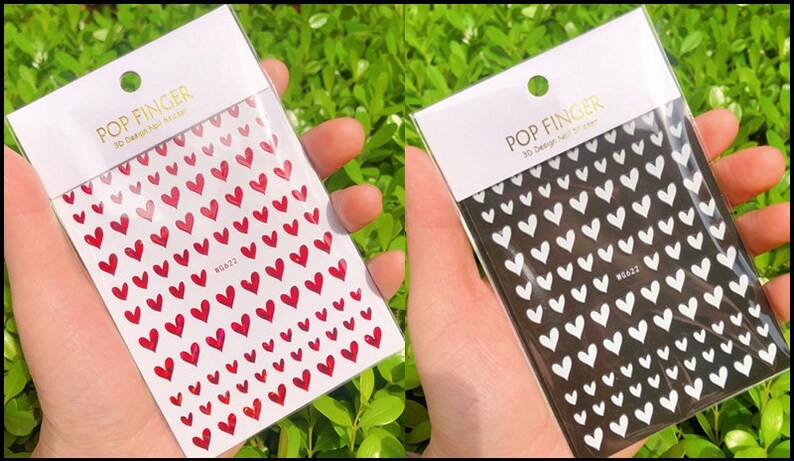 Op de afbeelding: Twee verpakkingen met 3D nagel stickers met een hartjes ontwerp. E&eacute;n verpakking heeft rode hartjes op een witte achtergrond en de andere heeft witte hartjes op een zwarte achtergrond. De verpakkingen zijn gelabeld met "POP FINGER 3D Design Nail Sticker".