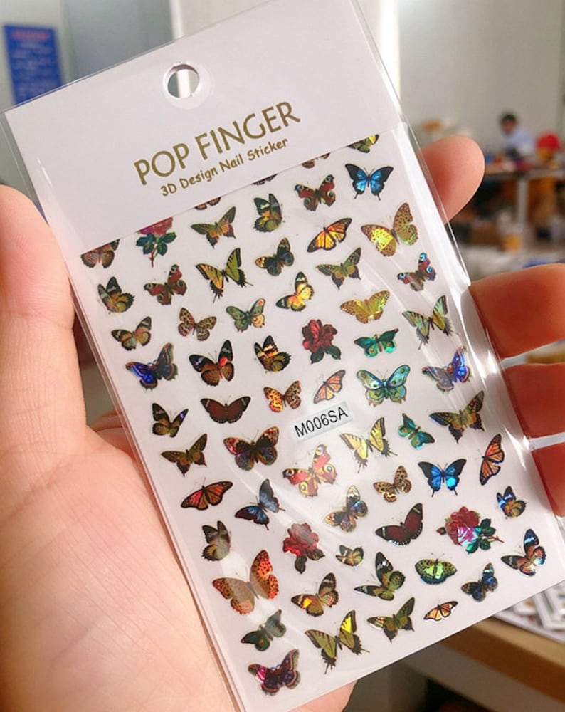 May include: A sheet of nail stickers with a variety of colorful butterfly designs. The stickers are 3D and have a holographic finish. The sheet is labeled "POP FINGER 3D Design Nail Sticker" and "M006SA".