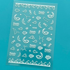 May include: A sheet of white nail decals featuring a moon, stars, clouds, and constellations design. The sheet is labelled "Moon Patterns" and "A048".