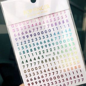 May include: A sheet of rainbow holographic nail stickers with numbers 0-9. The stickers are arranged in rows and columns, with each number appearing multiple times. The stickers are packaged in a clear plastic bag with the brand name "POP FINGER" and the text "25 Design Nail Stickers" printed on the front.