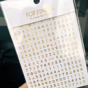 May include: Gold number nail stickers on a white background. The numbers range from 0 to 9 and are arranged in a grid pattern. The stickers are designed for nail art and are labeled "POP FINGER 3D Design Nail Sticker".