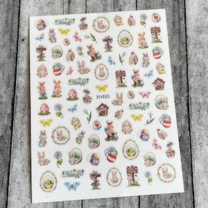 以下が含まれることがあります： ウサギ、卵、花、十字架など、さまざまなデザインが施されたイースターネイルステッカーのシート。ステッカーは白い背景に印刷されており、パステルカラーが特徴です。シートには、「イースター祝福」と「XH495」のテキストも含まれています。