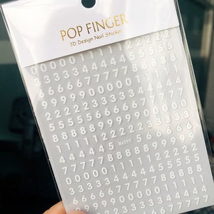 May include: A sheet of white nail stickers with numbers 0-9 printed on them. The stickers are arranged in rows and columns. The sheet is labeled "POP FINGER 3D Design Nail Sticker" and "Me099".