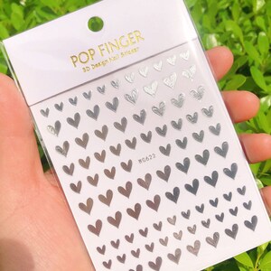 Op de afbeelding: Een vel zilveren nagelstickers met een ontwerp van kleine hartjes. Het vel is gelabeld met "POP FINGER 3D Design Nail Sticker" en "WG622".