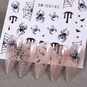 May include: A sheet of nail art stickers featuring spiders, spiderwebs, and other Halloween-themed designs. The stickers are black and white, with blue accents. Five long, glittery pink nails are decorated with the stickers. The sheet is labeled "SW-CS142".