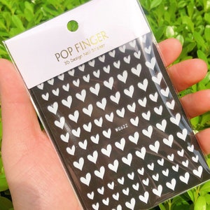 Op de afbeelding: Een vel met witte hartvormige nagelstickers op een zwarte achtergrond. Het stickerblad is gelabeld met "POP FINGER 3D Design Nail Sticker" en "WG622".