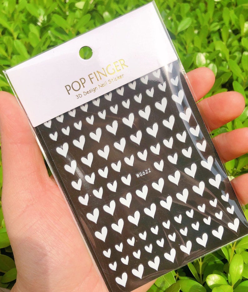 Op de afbeelding: Een vel met witte hartvormige nagelstickers op een zwarte achtergrond. Het stickerblad is gelabeld met "POP FINGER 3D Design Nail Sticker" en "WG622".