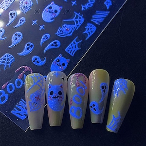 May include: Halloween-themed nail art featuring blue glowing designs of ghosts, cats, and spiderwebs. The nails are painted with a light yellow base and accented with glitter. A sheet of matching nail stickers is visible.