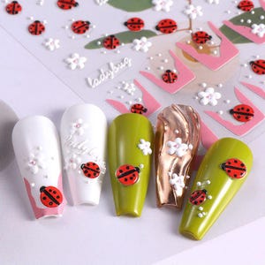 May include: Nail art stickers featuring ladybug and flower designs. The stickers include red ladybugs with black spots and white flowers. The nails are painted white, olive green, and gold. The word "ladybug" is written on the sticker sheet.