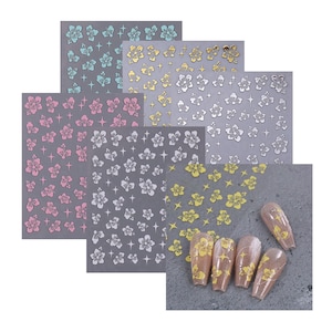 May include: Set of five sheets of nail art stickers featuring a floral design. The stickers are in various colors, including gold, silver, pink, and blue. The stickers are shown on a gray background with a hand model wearing the stickers on their nails.