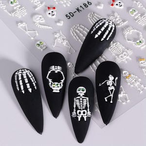 May include: Black nail art with white skeleton designs. The nails feature various skeleton-themed decorations, including hands, skulls, and full skeletons. The nail art is set against a clear background with additional skeleton stickers. The text "5D-K186" is visible.