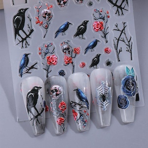 May include: Sheet of nail art stickers featuring crows, skulls, red roses, and spiderwebs. The designs include black crows perched on branches, skulls with red roses, and blue crows. The nail art stickers are on a clear background.