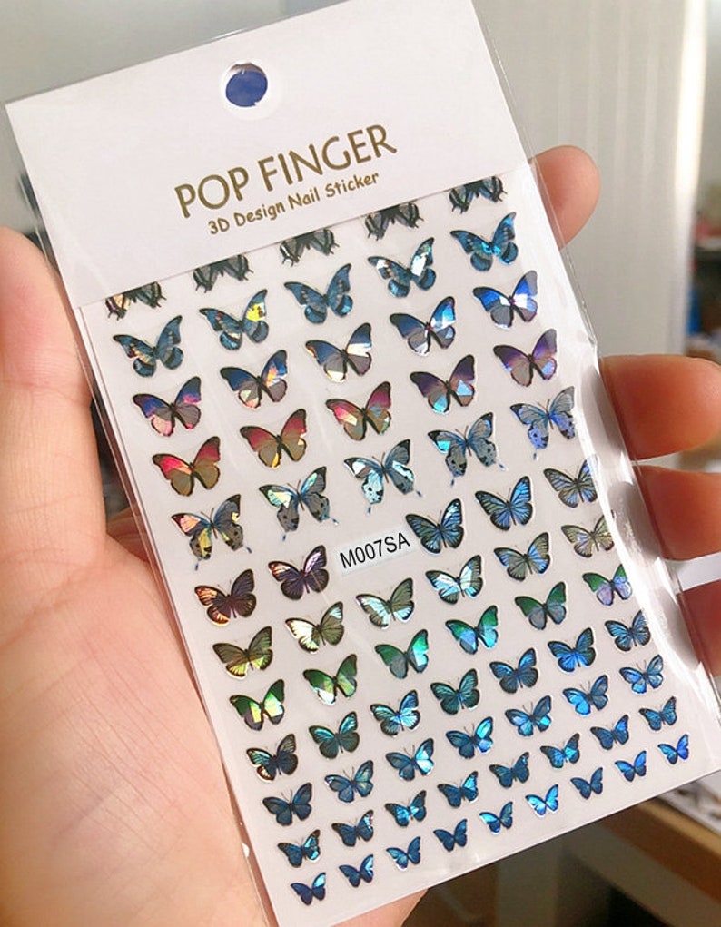 May include: A sheet of iridescent butterfly nail stickers. The stickers are arranged in rows and feature a variety of blue, green, and purple butterflies. The sheet is labeled "POP FINGER 3D Design Nail Sticker" and "M007SA".