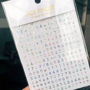 May include: A sheet of iridescent holographic nail stickers with numbers 0-9. The stickers are on a white background with the text "POP FINGER 3D Design Nail Sticker" and "Me099" printed in gold.