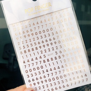 May include: A sheet of gold-colored number stickers for nail art. The numbers 0 through 9 are arranged in rows on a white background. The sheet is labeled "POP FINGER 3D Design Nail Sticker" and "Me099".