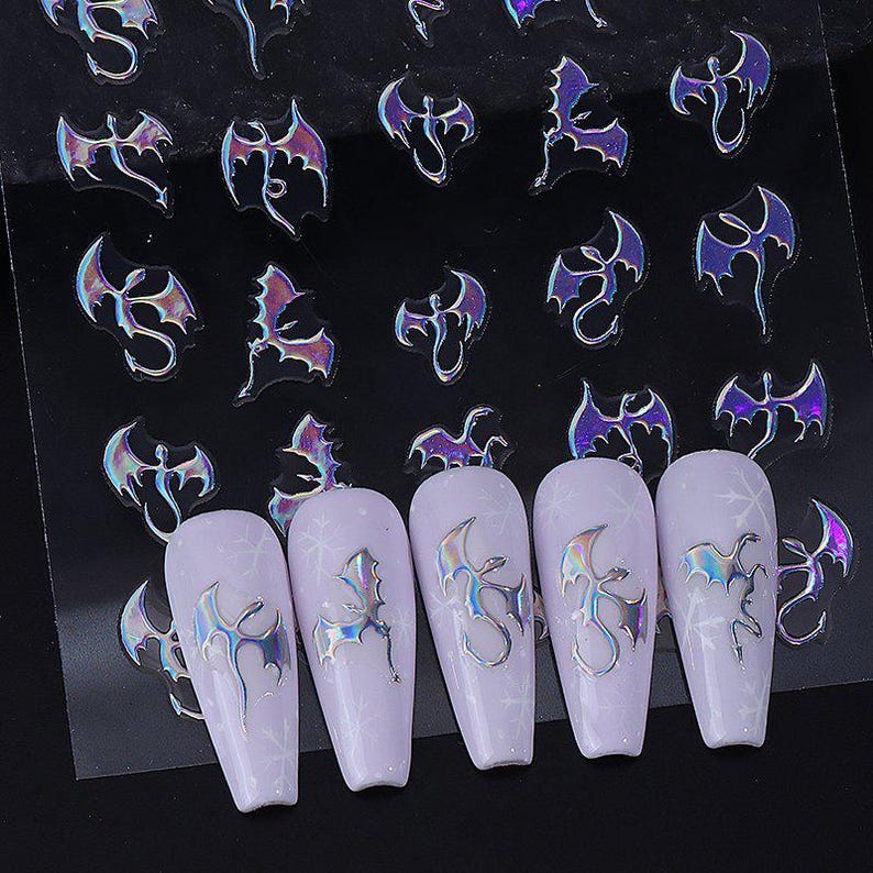 May include: Sheet of iridescent bat nail art stickers. The stickers are in various sizes and feature a bat design with purple and silver holographic effects. Several nails are decorated with the bat stickers, showcasing the nail art.
