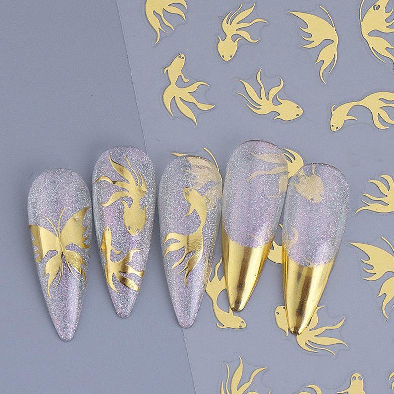 May include: A set of five artificial nails with a glittery silver base and gold foil designs. The designs include a butterfly, fish, and gold tips. Additional gold fish designs are scattered around the nails.