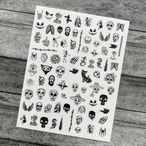 May include: A sheet of black and white temporary tattoos featuring various spooky designs. The sheet includes skulls, bats, cats, wings, and other Halloween-themed illustrations. The tattoos are arranged on a white background.