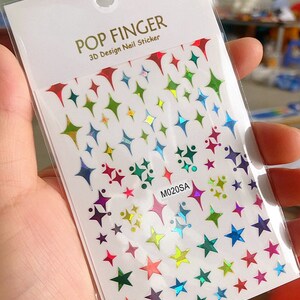 May include: A sheet of rainbow iridescent star and diamond shaped nail stickers. The sheet is white with the text "POP FINGER 3D Design Nail Sticker" and "M020SA" printed on it.