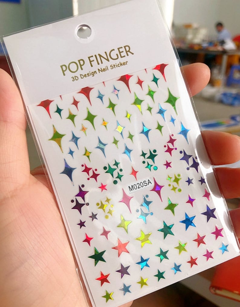 May include: A sheet of rainbow iridescent star and diamond shaped nail stickers. The sheet is white with the text "POP FINGER 3D Design Nail Sticker" and "M020SA" printed on it.