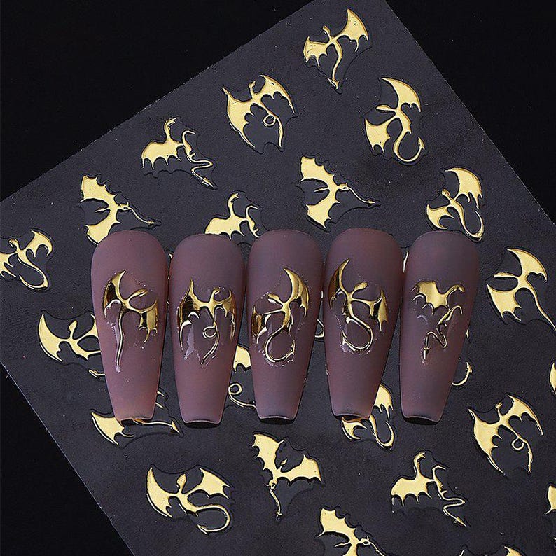 May include: A sheet of gold-colored dragon nail art stickers on a black background. Five matte mauve-colored artificial nails are adorned with the same dragon design. The stickers are a metallic gold color.