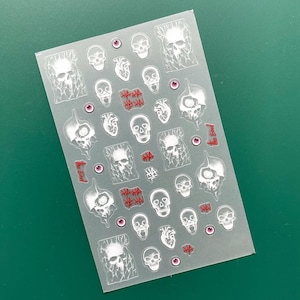 Może przedstawiać: Przezroczysty arkusz naklejek z różnymi wzorami czaszek, sercami, gałkami ocznymi oraz tekstem "HA HA HA!" i "The End" w kolorze czerwonym. Czaszki są w różnych stylach, niektóre ze spękanymi detalami.