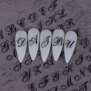 Könnte beinhalten: Nagelkunst-Aufkleber mit schwarzen Schreibschriftbuchstaben. Fünf weiße Nägel zeigen die Buchstaben D, A, J, B und U. Der Hintergrund ist eine transparente Folie mit verschiedenen Buchstabenmustern.
