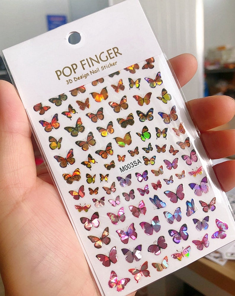 May include: A sheet of iridescent butterfly nail stickers. The stickers are in various shades of pink, purple, orange, and yellow. The sheet is labeled "POP FINGER 3D Design Nail Sticker" and "M003SA".
