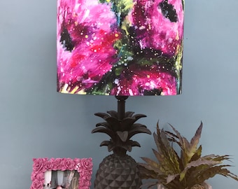 May include: A black pineapple-shaped lamp base with a pink and green floral lampshade. The lampshade has a watercolour-like design with abstract flowers and splatters of paint.