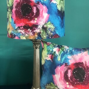 May include: A silver lamp with a blue and pink floral lampshade. The lampshade features a large pink flower with a black centre. The lamp is on a table with a matching cushion.