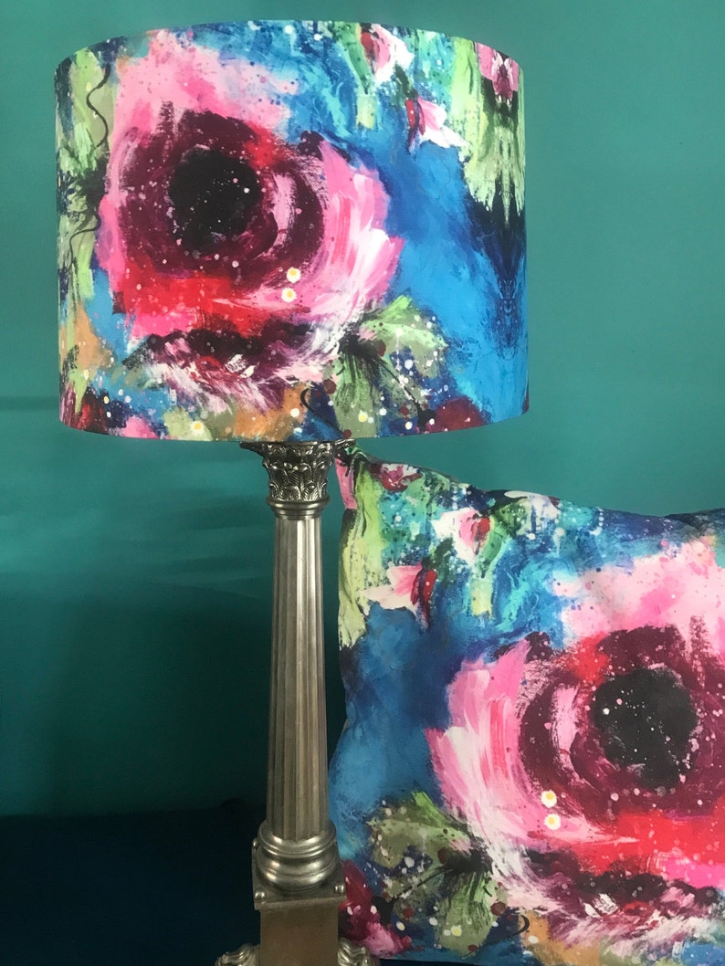 May include: A silver lamp with a blue and pink floral lampshade. The lampshade features a large pink flower with a black centre. The lamp is on a table with a matching cushion.