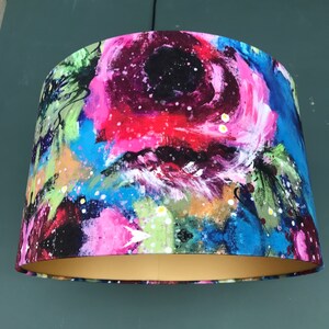 May include: A colourful lampshade with a floral design. The lampshade is made of fabric and has a white, pink, blue, green and black abstract floral pattern. The lampshade has a gold trim around the bottom edge.