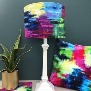May include: A white table lamp with a colourful abstract print lampshade. The lampshade has shades of blue, green, yellow, pink, and purple. The lamp is on a white base with a square shape.