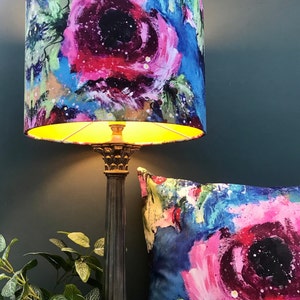 May include: A lampshade with a floral pattern in shades of pink, blue, and green. The lampshade is lit and the light shines through the fabric. The lampshade is on a black lamp base.